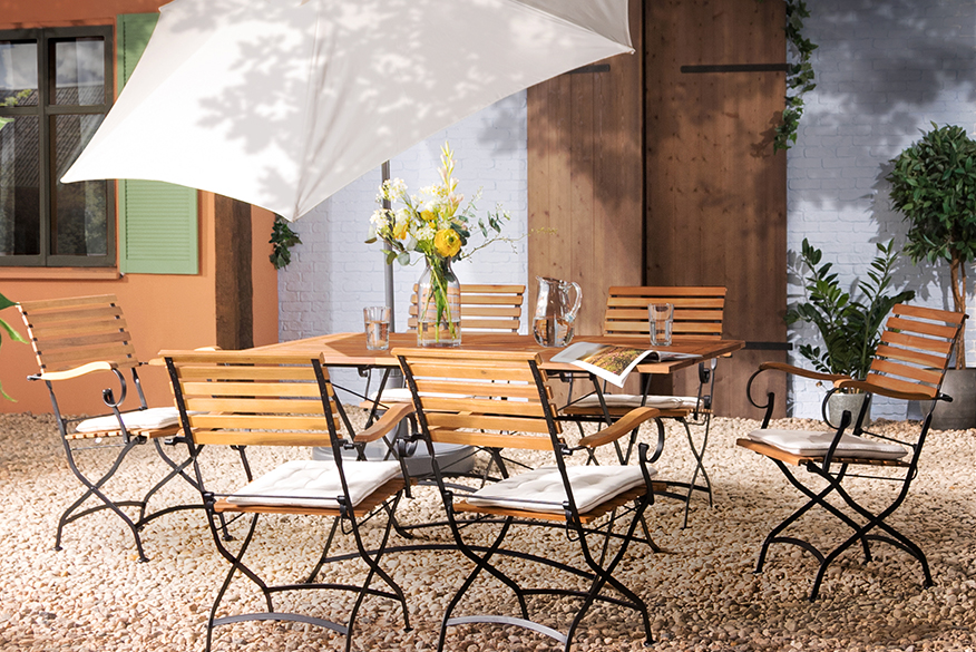 Terrasse mit Gartentisch, Gartenstühlen und Gartenbank aus Akazienholz mit hellblauen Sitzkissen, dazu ein weißer Sonnenschirm, ein gemusterter Outdoorteppich und Deko.