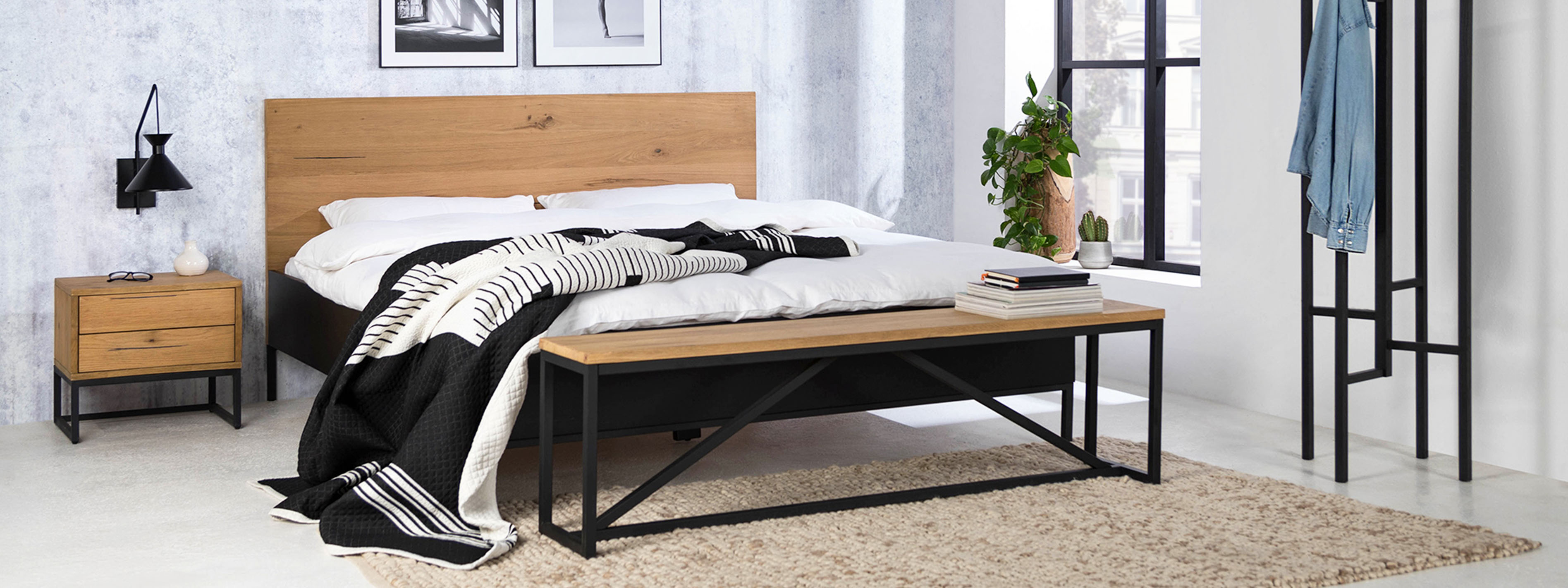 Schlafzimmer im minimalistischen Stil mit Bett, Nachttisch und Sitzbank der Serie Flox im Industrial-Stil aus Eichenholz und schwarzem Metall. Dazu schwarz-weiße Wandbilder, eine schwarze Wandleuchte, ein beiger Teppich und ein schwarzer Garderobenständer aus Metall.