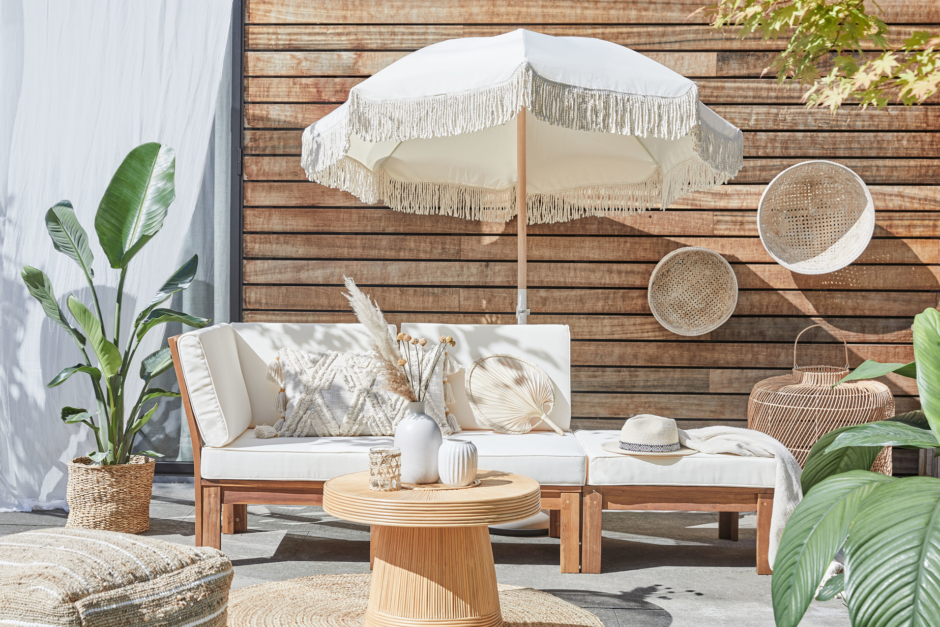 Outdoor-Loungemöbel der Serie LEXI by BUTLERS aus hellem Akazienholz mit weißen Polstern, dazu ein passender Tisch dekoriert mit weißen Vasen, ein weißer Sonnenschirm mit Fransen, ein Outdoor-Teppich und Pouf aus Jute sowie Wanddeko, Pflanzenkorb und Windlicht in Flechtoptik.