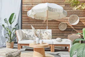 Outdoor-Loungemöbel der Serie LEXI by BUTLERS aus hellem Akazienholz mit weißen Polstern, dazu ein passender Tisch dekoriert mit weißen Vasen, ein weißer Sonnenschirm mit Fransen, ein Outdoor-Teppich und Pouf aus Jute sowie Wanddeko, Pflanzenkorb und Windlicht in Flechtoptik.