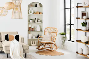 Fauteuil à bascule en rotin à côté d'un tapis rond en jute. Avec un canapé d'angle beige de la marque kollected, trois lampes suspendues en rotin, des coussins et un plaid ainsi que des vases, des chandeliers et des livres pour la décoration.