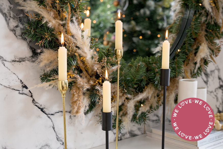 Weiße Kerzen in goldenen und schwarzen Kerzenhaltern vor einem Weihnachtskranz an einer Marmorwand