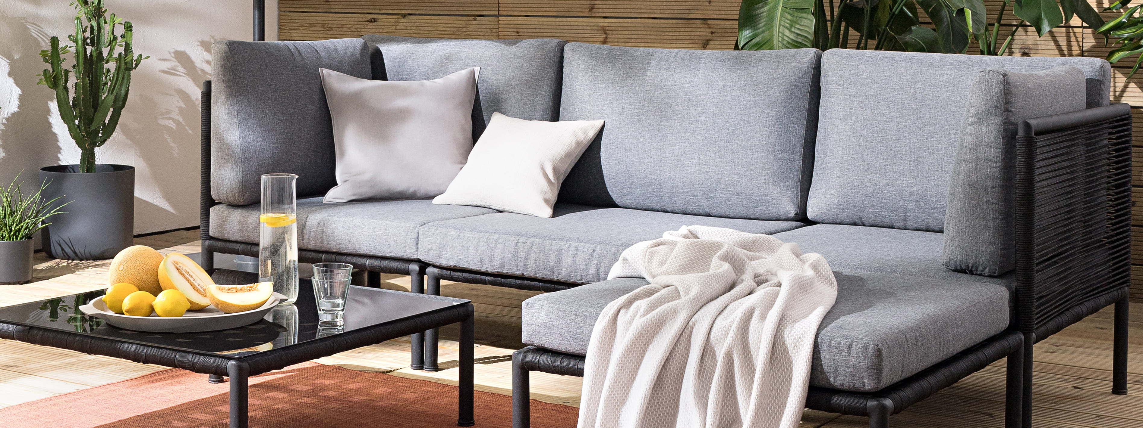 Outdoor-Loungesofa mit grauen Polstern, Kissen und Plaid, dazu ein schwarzer Tisch, Outdoor-Teppich und Grünpflanzen