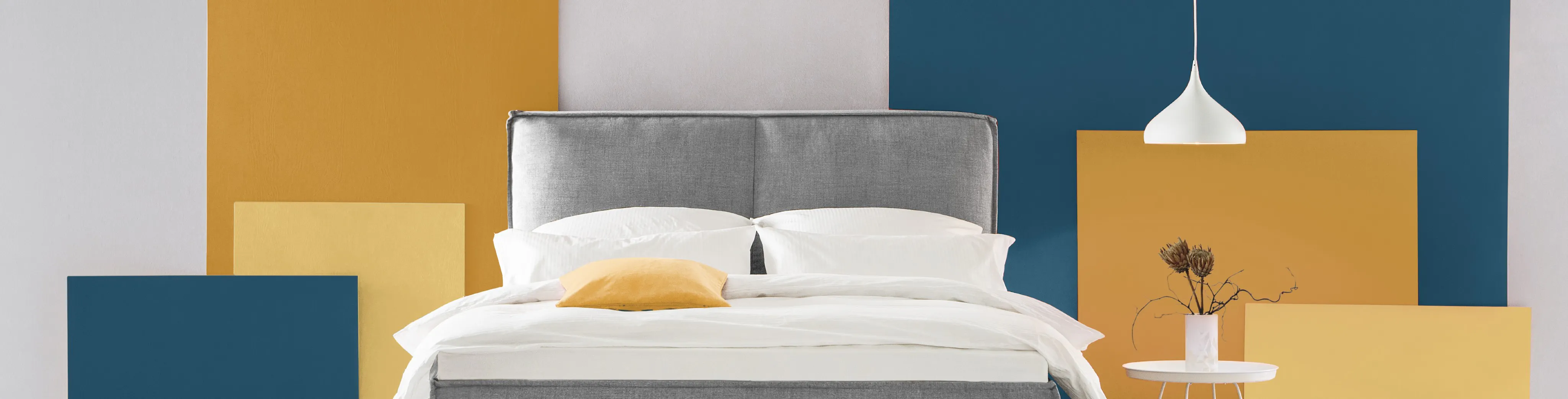Lit capitonné gris, avec derrière le lit, un mur jaune moutarde, blanc et bleu sombre, une lampe blanche et une petite table de chevet blanche.