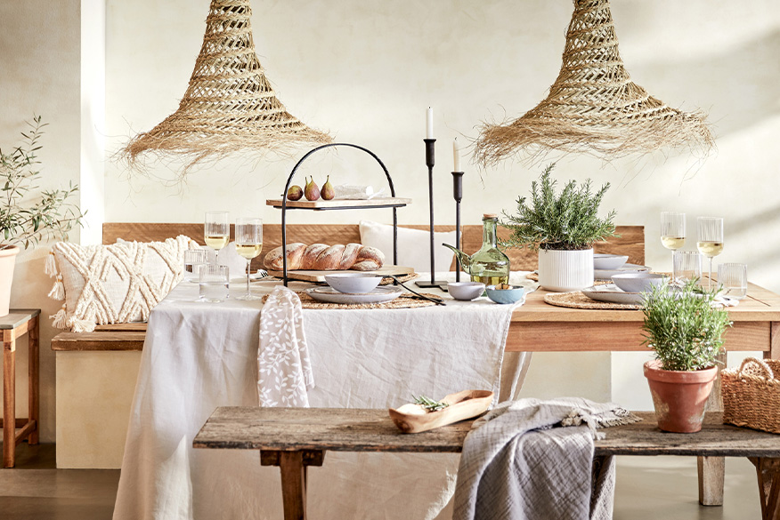 Esszimmer mit Produkten von BUTLERS im mediterranen Look: ein dekorierter Holztisch mit einer weißen Tischdecke, einer Etagere und Kerzenständern in Schwarz, Vasen, Geschirr und Gläsern; dazu zwei Pendelleuchten aus Rattan, eine rustikale Holzbank mit Körbchen, Plaid und Kräutern, ein Beistelltisch mit einem Pflanzenkübel sowie Kissen mit Fransen.