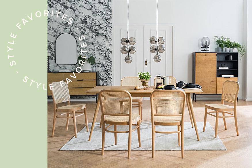 Heller Holztisch mit sechs Esszimmerstühlen mit Wiener Geflecht in einem Altbau-Esszimmer mit Holzboden, weißen Wänden, Kugellampen und Kommoden im Hintergrund, wobei die linke vor einer schwarz-weiß marmorierten Tapete steht