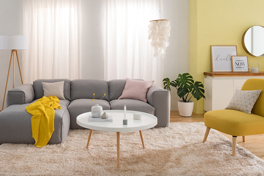 Salon avec canapé gris clair et fauteuil jaune, tous deux accessoirisés de plaid et coussins, table basse blanche et lampadaire scandinave, suspension en nacre, buffet blanc.