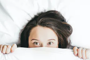 Junges Mädchen, das mit weit aufgerissenen Augen unter einer weißen Bettdecke hervorschaut