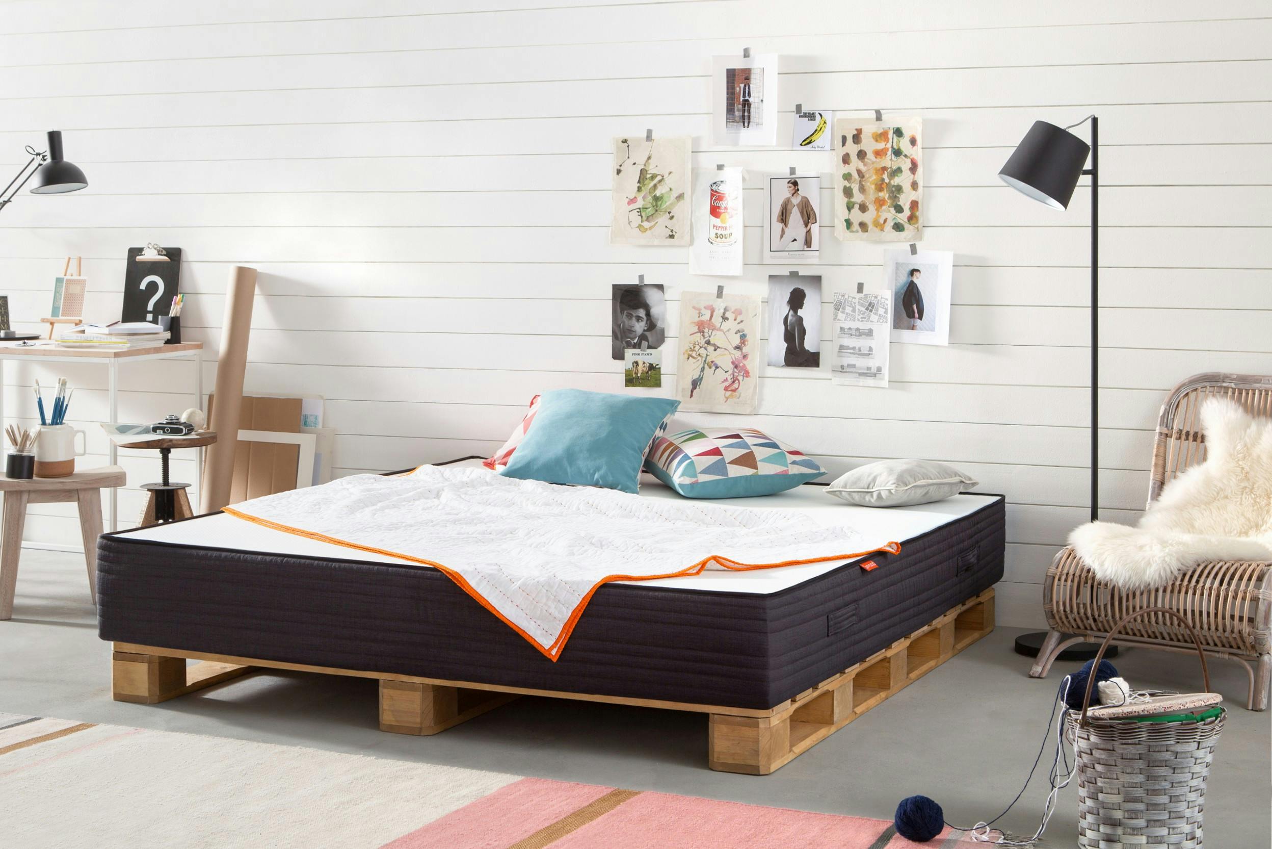 Zimmer mit Schreibtisch, Stehlampe, Bildern an der Wand und einem Palettenbett mit Matratze