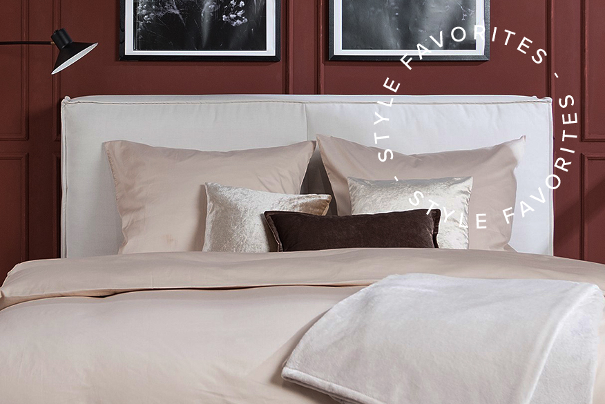 Schlafzimmer mit schokobrauner Wandfarbe und beigem Teppich, auf dem ein weißes Polsterbett mit cremefarbener Bettwäsche und schokobraunem Samtkissen steht; daneben ein Bild von Schokoladentafeln, die als Farbinspiration zum Einrichten mit Braun dienen.