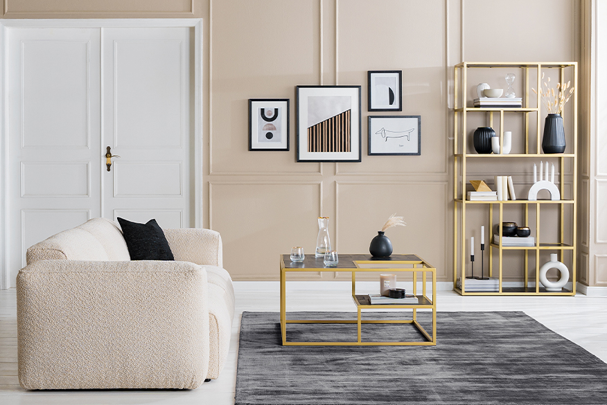 Salon avec canapé et fauteuil beiges, table basse, étagère et enfilade noires avec structure en métal doré, objets décoratifs blancs, tapis et coussins.