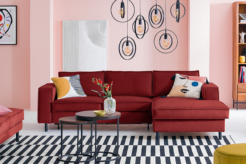 Rotes Sofa inkl. Polsterhocker der home24 Exklusivserie Fort Dodge by kollected mit bunten Kissen auf einem schwarz-weiß gestreiften Teppich. Dazu zwei runde Beistelltische aus dunklem Holz und Metall im Industrial-Stil, ein gelber Pouf, ein schwarzer Stuhl mit Wiener Geflecht und gelbem Kissen, mehrere schwarze Pendelleuchten, ein Kandinsky-Wandbild und Highboard Deva aus Holz und Metall.