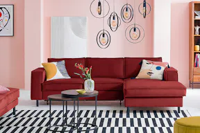Canapé rouge avec repose-pieds Fort Dodge, de la marque exclusive home24 kollected, avec des coussins colorés, sur un tapis rayé noir et blanc. En sus, deux tables basses rondes en bois foncé et métal dans un style industriel, un pouf jaune, une chaise noir en cannage avec un coussin jaune, plusieurs suspensions noires, une affiche de Kandinsky et une commode Deva en bois et métal.