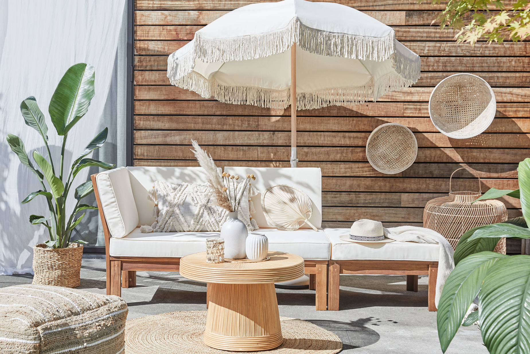 Outdoor-Loungemöbel der Serie LEXI by BUTLERS aus hellem Akazienholz mit weißen Polstern, dazu ein passender Tisch dekoriert mit weißen Vasen, ein weißer Sonnenschirm mit Fransen, ein Outdoor-Teppich und Pouf aus Jute sowie Wanddeko, Pflanzenkorb und Windlicht in Flechtoptik.