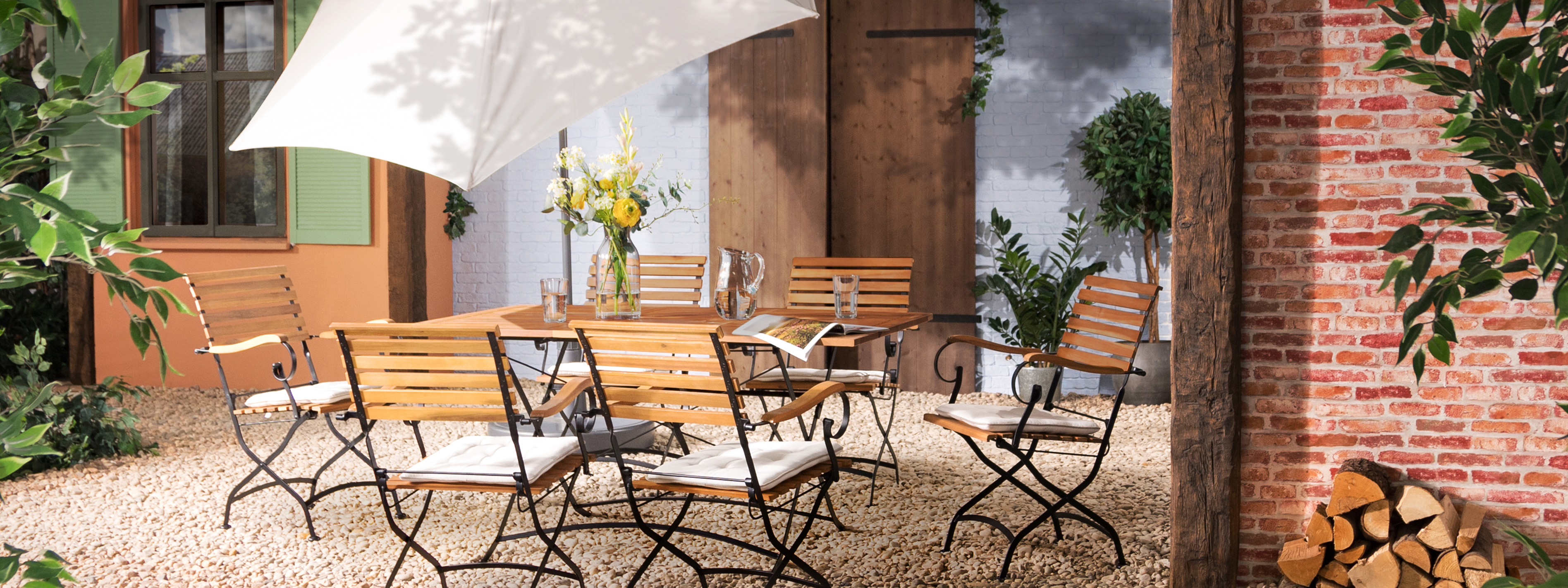 Terrasse mit Gartentisch, Gartenstühlen und Gartenbank aus Akazienholz mit hellblauen Sitzkissen, dazu ein weißer Sonnenschirm, ein gemusterter Outdoor-Teppich und Deko.
