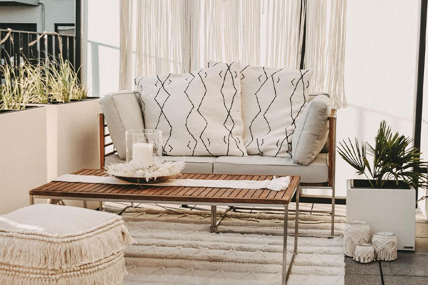 Terrasse im Boho-Stil mit Outdoorloungemöbeln aus hochwertigem Akazienholz, hellen Textilien in Cremeweiß wie Kissen, Teppich und Sitzpouf mit Fransendetails sowie eine Makramee-Girlande.