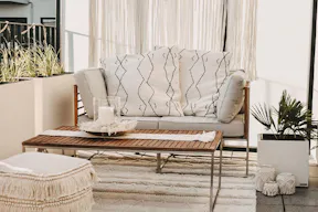 Terrasse im Boho-Stil mit Outdoorloungemöbeln aus hochwertigem Akazienholz, hellen Textilien in Cremeweiss wie Kissen, Teppich und Sitzpouf mit Fransendetails sowie eine Makramee-Girlande.