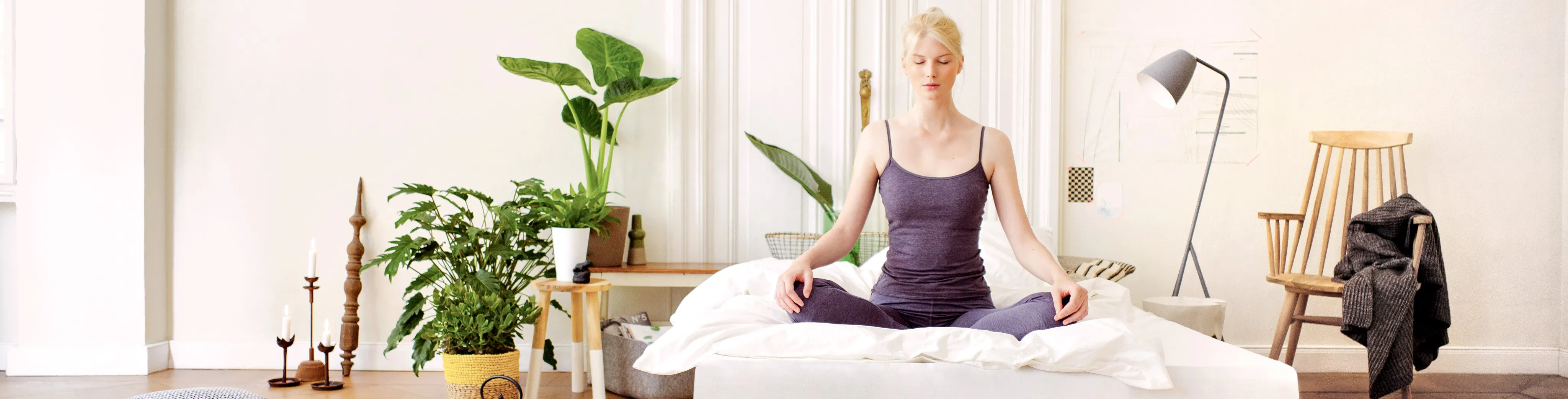 Frau im Schneidersitz auf dem Bett in einem Zimmer mit Grünpflanzen und grauer Stehlampe