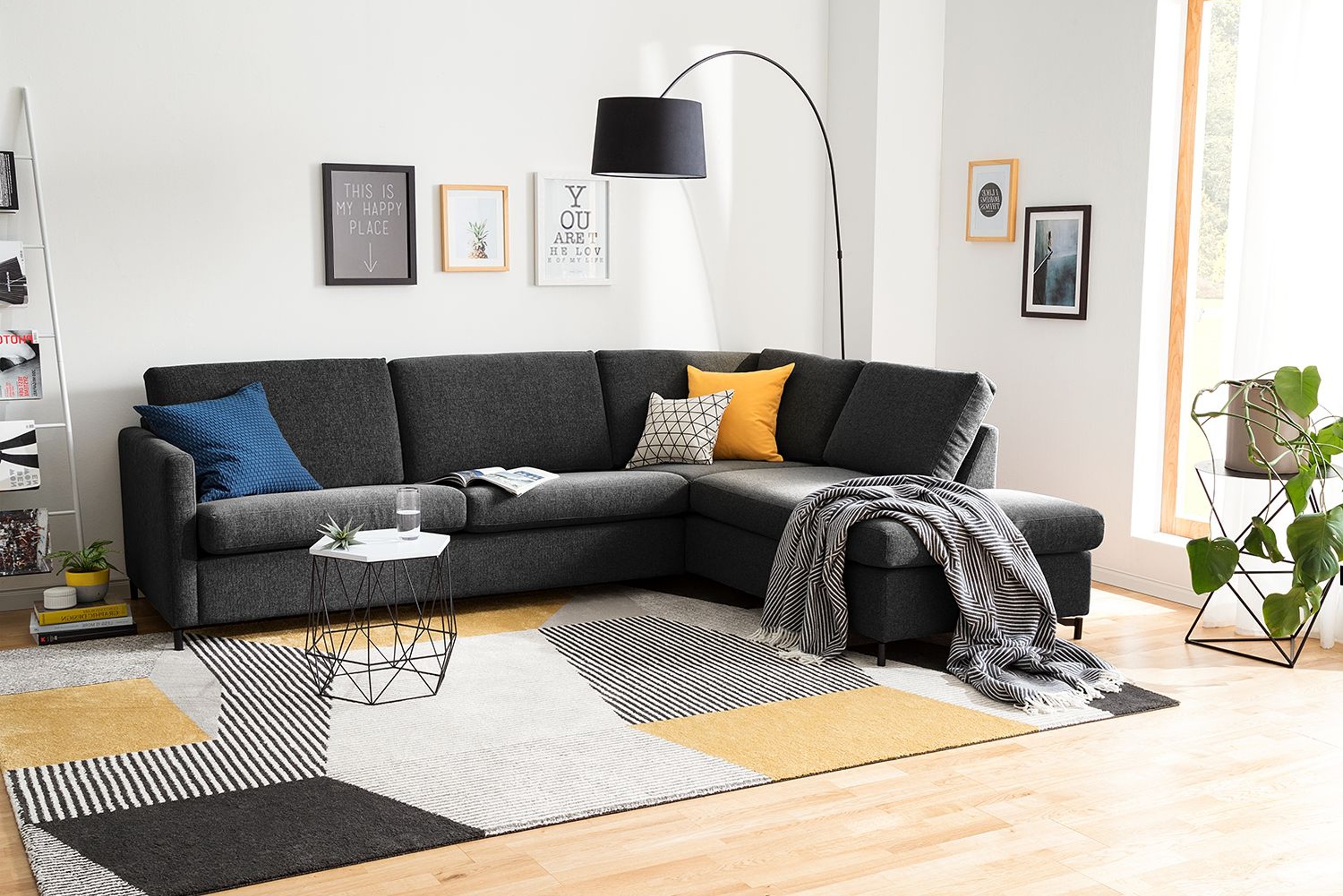 Zwarte bank met zwarte staande booglamp en gekleurde sierkussens met daarvoor kleine, geometrische bijzettafel en geometrisch vloerkleed in wit, wart en oker.