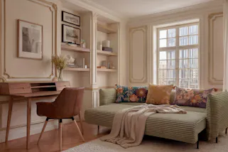 Ausklappbares Schlafsofa aus hellgrünem Cord mit bunten Blümchen-Kissen im Boho-Look, dazu ein Sekretär aus dunklem Holz im Retro-Stil sowie ein Armlehnenstuhl aus dunklem Leder.