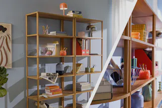 Modernes Standregal aus Holz vor himmelblauer Wand, dekoriert mit Körben, Büchern und Deko, dazu ein Holzregal mit Büchern und Schreibartikeln.