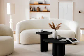 Wohnzimmer in organischen Formen mit Sessel und Sofa Osane in Creme sowie dem schwarzen Couchtisch Besare von Studio Copenhagen, dazu ein weißer Teppich, eine weiße Steh- und Tischleuchte, Keramik- und Holzdeko in Erdtönen sowie ein schwarzes Sideboard.