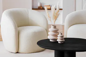 Le fauteuil Osane et la table basse Besare de la marque exclusive Studio Copenhagen de home24 sur un tapis beige, accompagnés de deux vases en terre cuite au design sculptural avec des tiges de pampas.