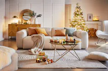 Un salon moderne et chaleureux décoré pour les fêtes de Noël. Au centre de la pièce, un grand canapé blanc est recouvert de coussins aux couleurs chaudes (orange, moutarde) et d'une couverture moelleuse. Devant le canapé, une table basse en verre repose sur un piétement métallique doré. Un tapis blanc recouvre le sol. À gauche, un meuble bas en bois clair abrite des objets décoratifs et une lampe de table dorée. À droite, un fauteuil à bascule blanc et un arbre de Noël illuminé apportent une touche festive à la scène. Sur le manteau de la cheminée, des bougies et des décorations complètent l'ambiance chaleureuse.