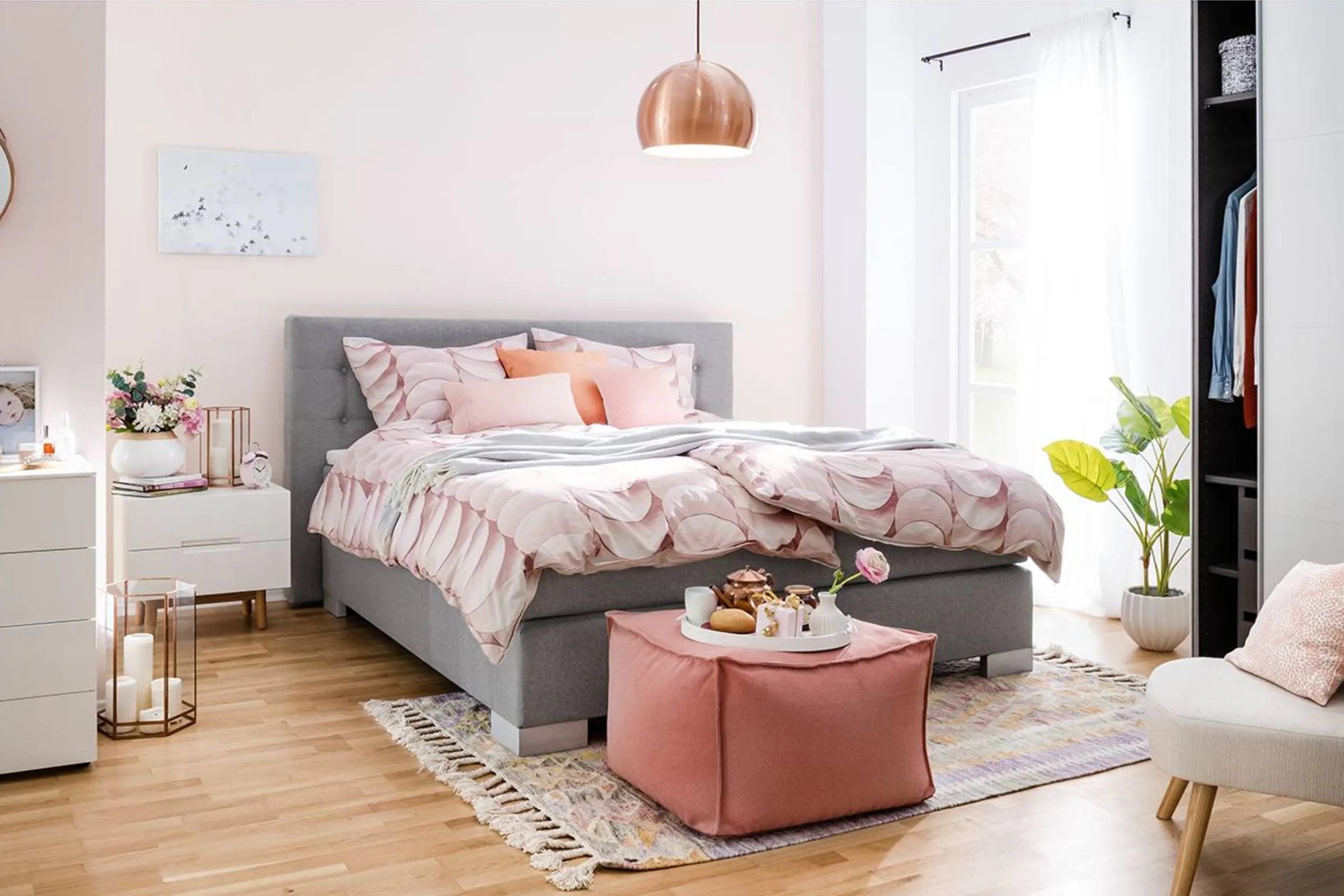 Schlafzimmer in Pastelltönen mit einem grauen Bett, rosa gemusterter Bettwäsche und einem Sitzpouf