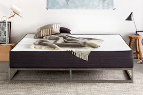 Dunkelblaue Matratze Smood mit weißer Liegefläche sowie beigen Kissen und einem beige kariertem Plaid auf einem Metallbett