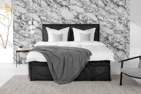 Boxspringbett KINX mit schwarzem Samtbezug vor einer auffälligen Marmorwand, darauf weiße Bettwäsche fürs Hotel-Feeling, kombiniert mit einer grauen Tagesdecke, davor ein beiger Wollteppich.