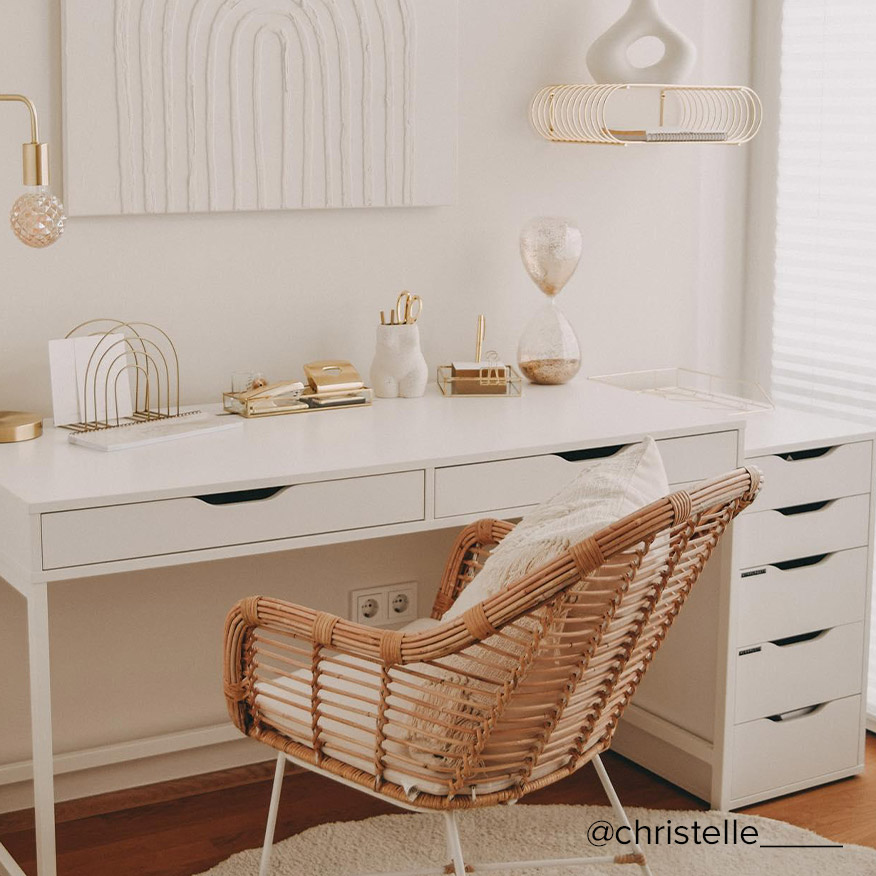Einfach und effektvoll eingerichtetes Homeoffice mit einem weißen Schreibtisch als Basis, dazu ein Rattanstuhl im Boho-Stil und reichlich goldene Wohnaccessoires für einen Touch Glamour, darüber ein weißes Strukturbild.