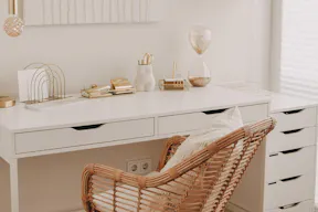 Einfach und effektvoll eingerichtetes Homeoffice mit einem weißen Schreibtisch als Basis, dazu ein Rattanstuhl im Boho-Stil und reichlich goldene Wohnaccessoires für einen Touch Glamour, darüber ein weißes Strukturbild.