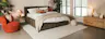 Schlafzimmer mit Polsterbett KINX der home24 Exklusivmarke Studio Copenhagen aus olivgrünem Cord, dazu Sessel Lovelock in Rostrot, Beistelltisch Cyra im Retro-Stil mit Wiener Geflecht, ein Sideboard aus Holz, Tischleuchten von BUTLERS, ein beiger Teppich, ein Pouf, Kissen und Decken in Erdfarben sowie Grünpflanzen.