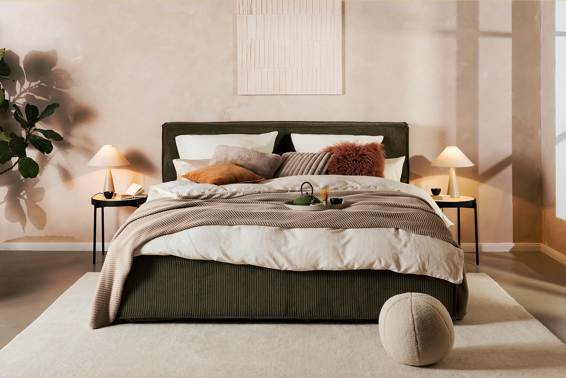 Polsterbett KINX von Studio Copenhagen aus olivgrünem Cord, dazu zwei Nachttische im Retro-Stil mit Wiener Geflecht, ein Sideboard aus Holz, Tischleuchten von BUTLERS, ein beiger Teppich, ein Pouf, weiße Bettwäsche, beige und dunkelrote Kissen, eine beige Tagesdecke, Grünpflanzen sowie ein Tablett mit Teekanne und Geschirr.