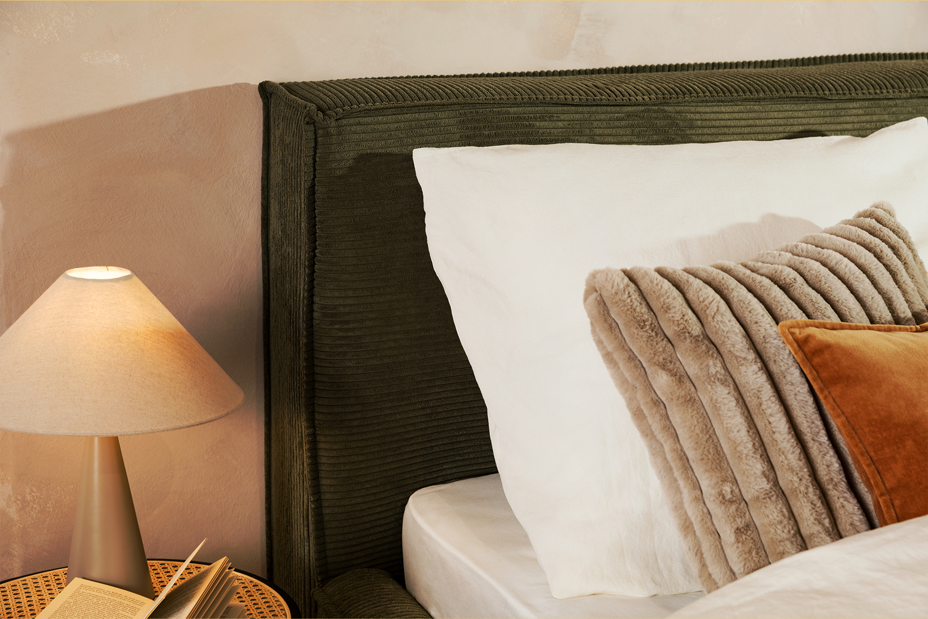Olivgrünes Polsterbett KINX im Boxspringbett-Design mit weißer Bettwäsche, einem beigen und rostroten Kissen, einem Nachttisch mit Wiener Geflecht sowie einer Tischlampe von BUTLERS.