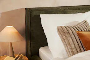 Olivgrünes Polsterbett KINX im Boxspringbett-Design mit weißer Bettwäsche, einem beigen und rostroten Kissen, einem Nachttisch mit Wiener Geflecht sowie einer Tischlampe von BUTLERS.