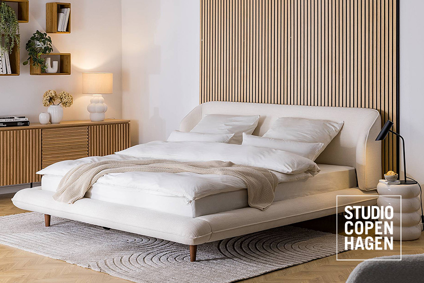 Polsterbett Kniss der home24 Designermarke Studio Copenhagen mit organisch geschwungenem Kopfteil und breitem Rahmen, bezogen mit beigem Webstoff.