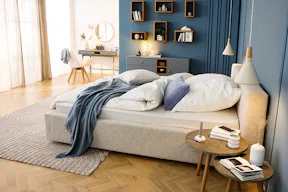Schlafzimmer in gedeckten Blau- und Grautönen mit einem grauen Polsterbett aus Boucléstoff, runden Nachttischen aus dunklem Holz, zwei weißen Pendelleuchten im Skandi-Stil, einem grauen Teppich, einem blauen Sideboard sowie Wandregalen aus Holz; dazu eine Schminkecke mit grauem Schminktisch, einem Stuhl im Skandi-Design sowie einem runden Wandspiegel.