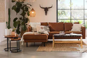 Wohnzimmer im Western-Style mit einer Fake-Ledercouch, lässigen Fransen an Kissen und Decken, einem Geweih als Wanddeko und vielen warmen Naturmaterialien und -tönen.