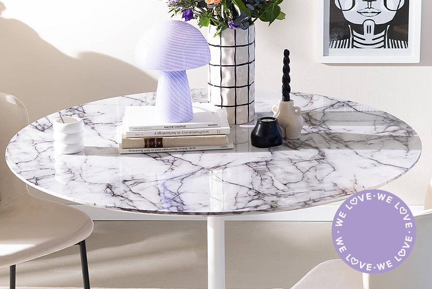 Runder Esstisch mit weißer Marmorplatte, beigen Polsterstühlen sowie farblich passender Deko wie einer schwarz-weißen Vase mit Frühlingsstrauß, lila Tischlampe im Mushroom-Look und schwarz-weißen Wandbildern.