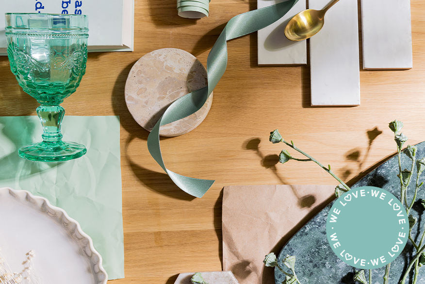 Salbeigrünes Gartenmöbel-Set bestehend aus zwei Metallbänken und einem Gartentisch aus Metall, welches auf einer Terrasse steht und mit Textilien in blaugrünen Aquatönen sowie Grünpflanzen dekoriert ist; daneben ein Moodboard mit verschiedenen Elementen wie einem türkisemen Glas, smaragdgrünem Marmortablett, salbeigrünem Papier und mehr, welches die Farbvielfalt von Aquatic Colors, also Meeresfarben, darstellen soll.