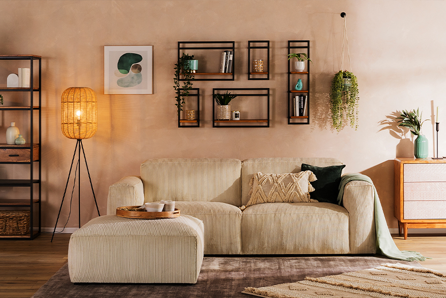 Gemütliches Boho-Wohnzimmer mit einem beigen Cordsofa, einer Holzkommode mit gemusterten Fronten, dazu viele Teppiche, Grünpflanzen, eine coole Stehlampe und Wandregale mit geometrischen Metallrahmen.