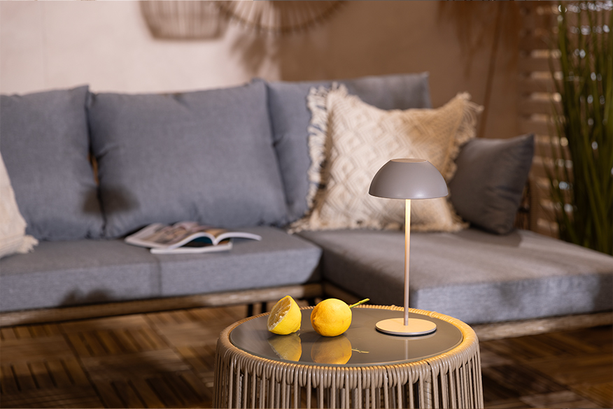 Graue, kleine Akku-Tischleuchte zum Mitnehmen für Garten oder Balkon, die angeknipst neben zwei Zitronenhälften auf einem Beistelltisch mit Rope Design vor einem grauen Outdoor-Loungesofa auf der Terrasse steht.