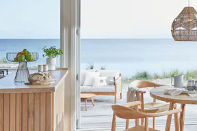 Offener Essbereich im Creme-Natur-Mix mit rundem Esstisch, Holzstühlen und Jute-Lampen. Ausserdem eine Terrasse mit einem Outdoorsofa aus Holz mit hellen Polstern sowie einem runden Beistelltisch.