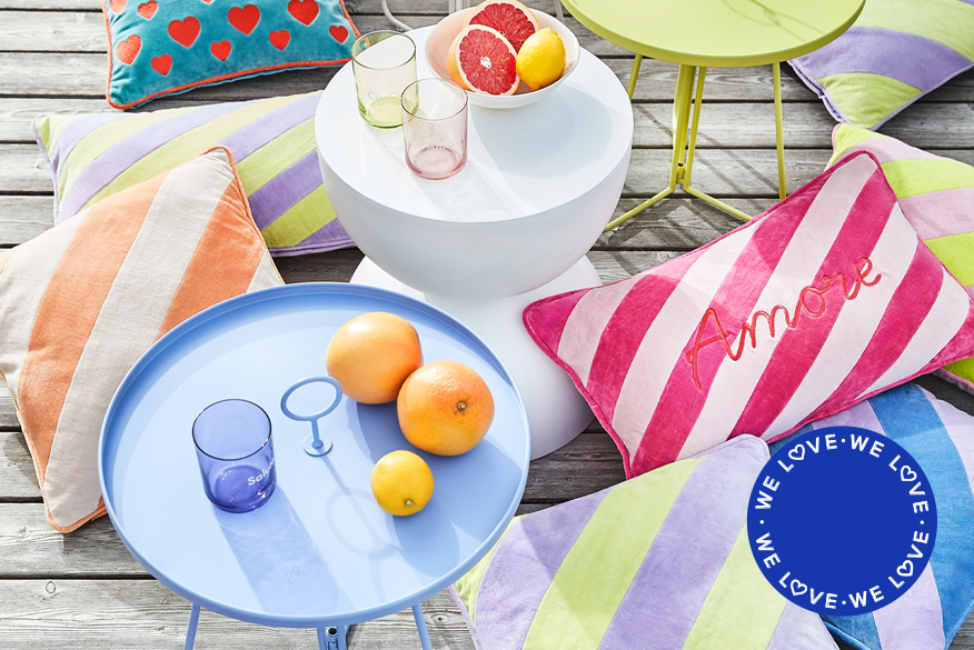 Verschiedene bunt gestreifte Kissen auf einer sonnigen Terrasse zusammen mit hellblauem und weißem Outdoor-Beistelltisch; daneben Gartenstühle mit Rückenlehnen aus angesagtem Rope Design in knalliger Flamingofarbe.