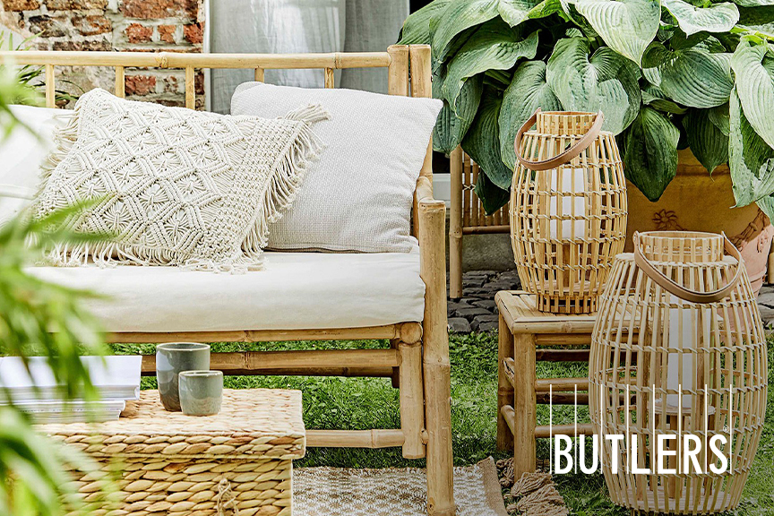 Zwei unterschiedlich große Laternen von BUTLERS aus Bambus, die im Boho-Garten neben einem Outdoor-Sofa auf der Wiese bzw. einem kleinen Beistelltisch stehen