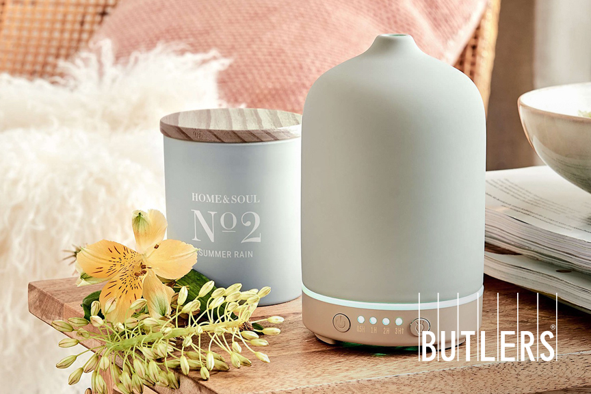 Aroma-Diffuser und Duftkerze von BUTLERS auf einer Holzbank, dekotiert mit Blüten sowie einem Schafsfell im Hintergrund.
