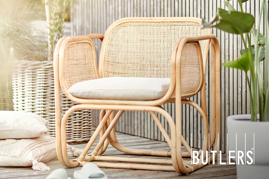Outdoor-Sessel bespannt mit natürlichem Rattangeflecht auf einer sonnigen Terrasse, dazu ein Jute-Teppich und Baumwollkissen mit Quasten und anderen Applikationen.