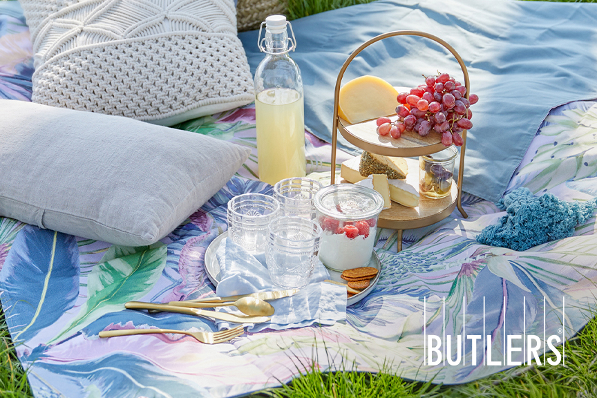 Blaue Picknickdecke im Grünen von BUTLERS mit Palmenprint, auf der graue Kissen, eine Etagère mit Käse, Quarkspeise mit Himbeeren und Saftgläser stehen.